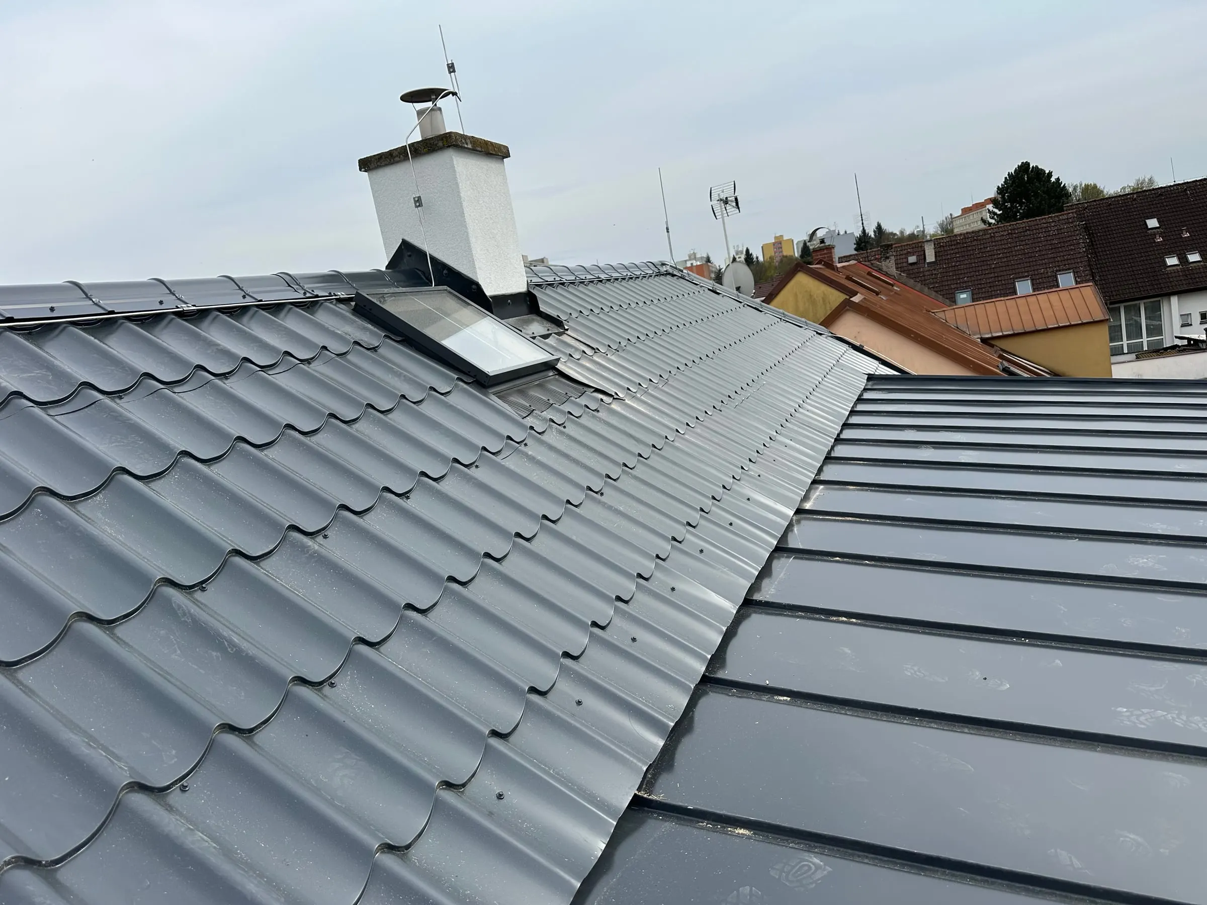 Detail střechy, kde plechová krytina v imitaci tašek navazuje na falcovaný plech, u komína střešní okno