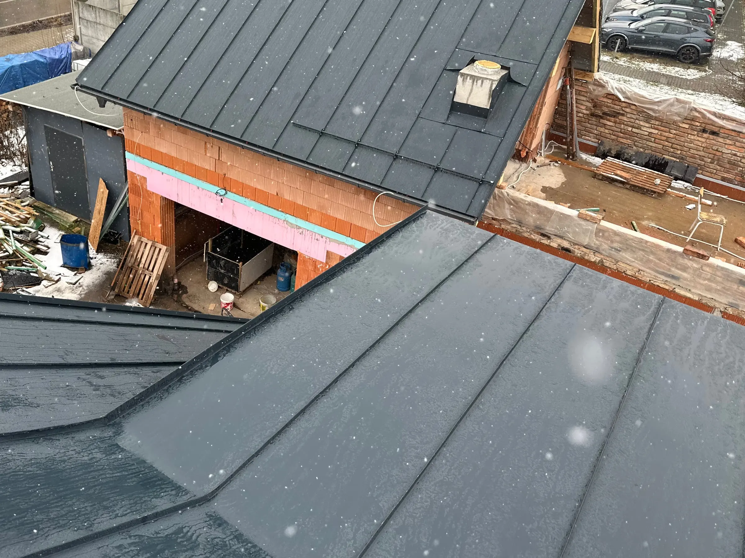 Pohled z hotové falcované střechy na přístavbu s novou tmavou falcovanou střechou, padá sníh